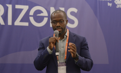 A person in a suit and glasses speaks into a microphone against a backdrop with the word 